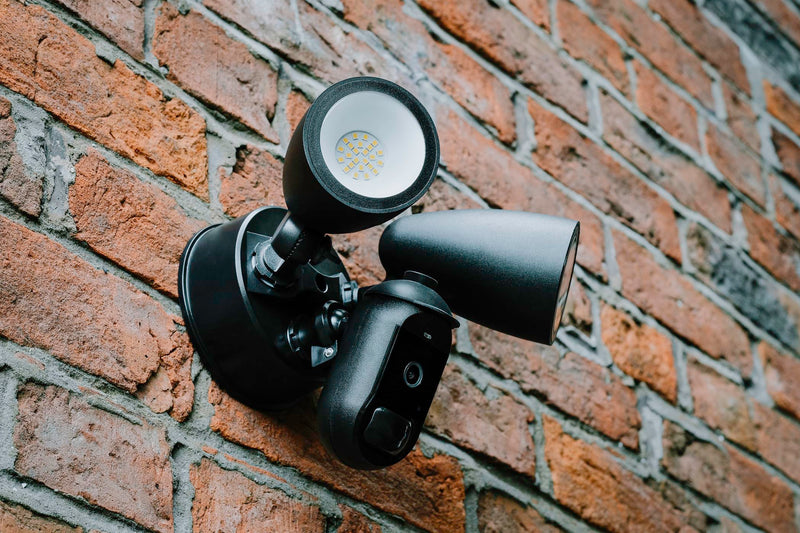 A black Amalock CAM-600FL-AC mains wired floodlight security camera mounted on a red brick wall