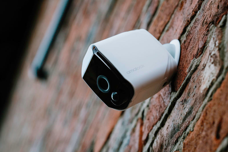 A white Amalock CAM200A wireless WiFi compact battery security camera branded with the Amalock logo mounted on a red brick wall