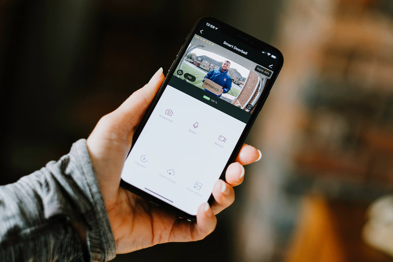 A hand holding a smartphone showing a livestream HD video from an Amalock DB101 wireless WiFi smart video doorbell 