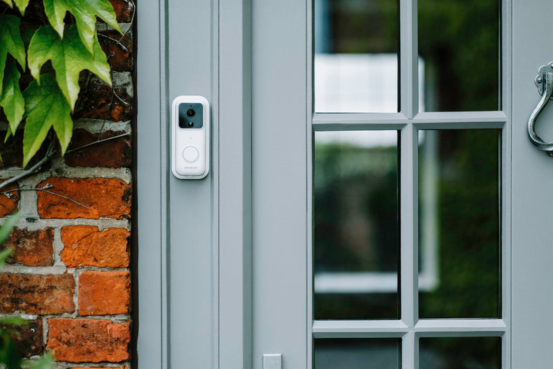 A white Amalock DB301 wireless video doorbell branded with the Amalock logo mounted on a grey doorframe