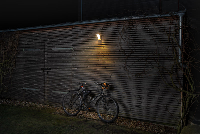 A white Ledvance Endura Pro Spot Sensor 8W cool white LED outdoor wall spotlight mounted on a garage wall with a bike leaning against it, illuminated at night. 