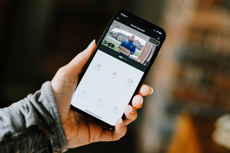 A hand holding a smartphone showing a livestream HD video from an Amalock DB101 wireless WiFi smart video doorbell 